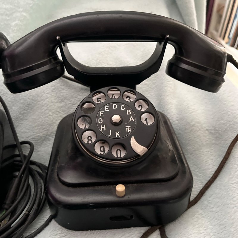 A vintage black rotary telephone with a circular dial featuring numbers, placed on a light surface. The handset rests on top of the base unit, and a cord is visible connecting to the device.
