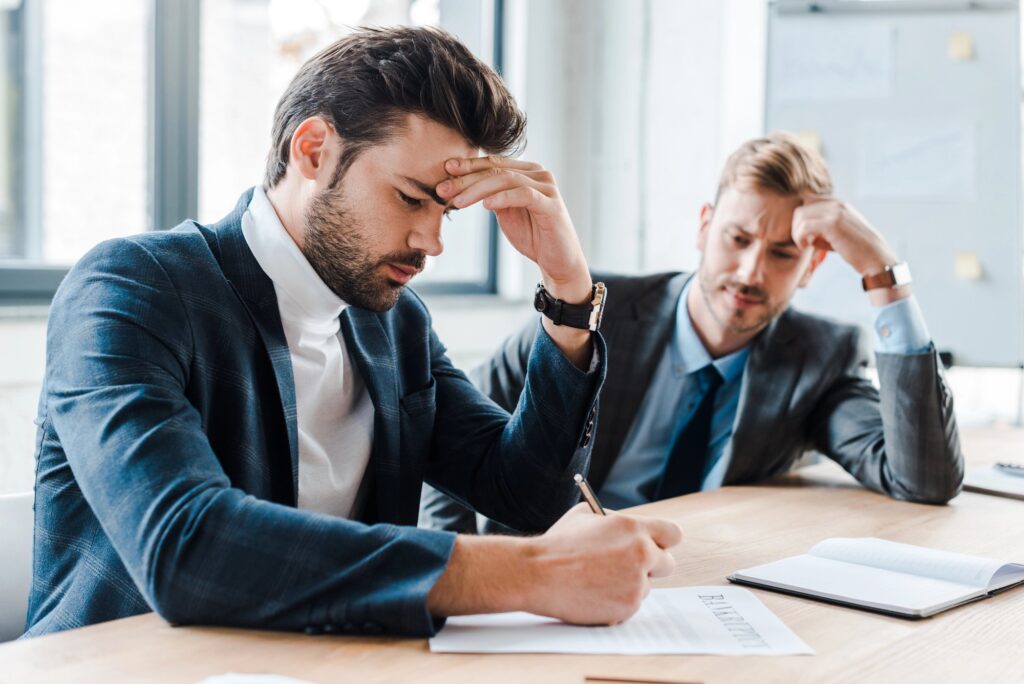Two businessmen in suits sitting at a table looking stressed and concerned while reviewing documents, representing the difficulty organizations face when confronting mistakes and failures.