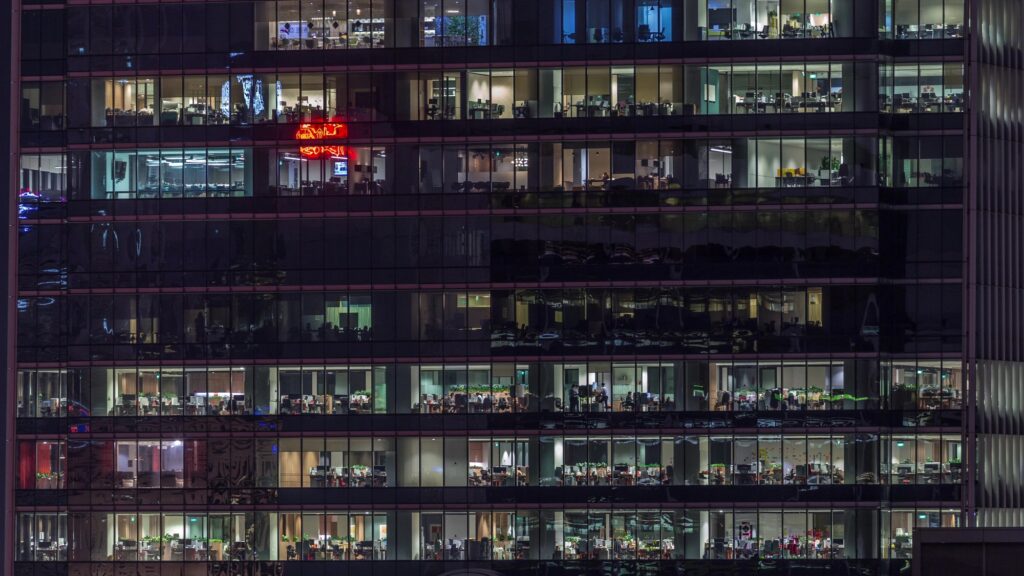 Modern office building at night with multiple illuminated floors showing different work activities across various levels. The transparent glass facade reveals people working in different spaces - meeting rooms, open work areas, and specialized departments - symbolizing the interconnected nature of knowledge management roles within an organizational structure. Warm interior lighting contrasts with the dark exterior, creating a visual metaphor for knowledge sharing across different organizational layers.