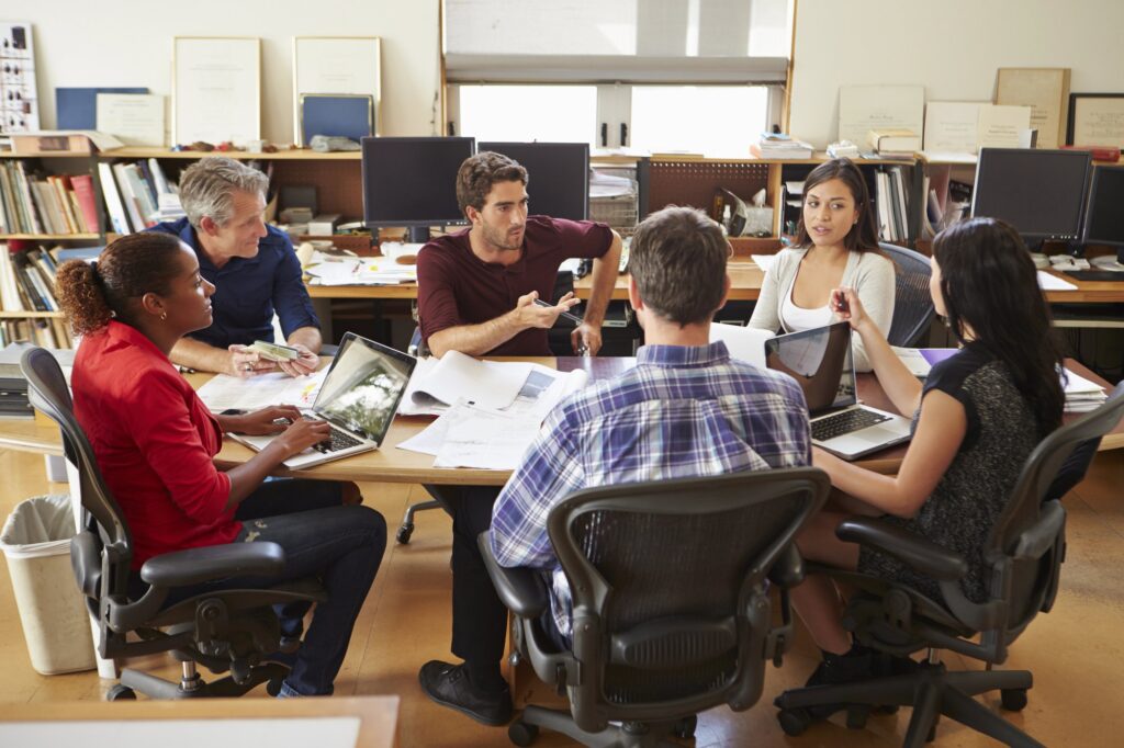 Six diverse professionals engaged in collaborative discussion around a wooden conference table in a modern office environment. The team members, representing different ages and backgrounds, sit in ergonomic chairs with laptops and documents, displaying open body language and active participation. Bookshelves and natural lighting in the background create a comfortable workspace atmosphere that promotes psychological safety and knowledge sharing. One team member gestures while explaining a point, while others listen attentively, embodying the trust and open communication essential for effective knowledge management.
