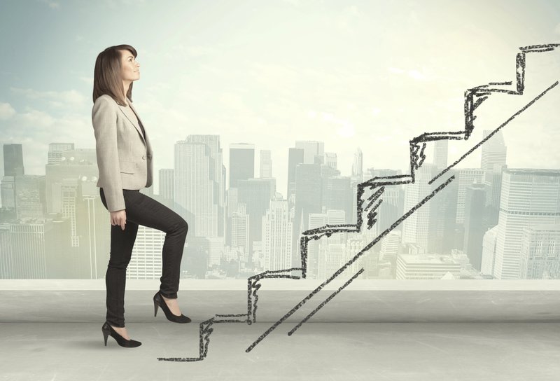 Professional businesswoman in beige blazer stepping up conceptual stairs drawn against city skyline background, symbolizing career advancement, skill development, and cognitive complexity progression in organizational learning