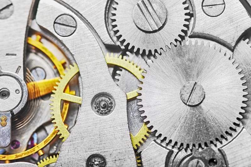 Close-up macro photograph of interlocking mechanical gears from a watch mechanism. Multiple metallic gears in brushed silver and steel mesh together precisely, with several golden brass gears creating visual accent. The image shows fine mechanical detail including gear teeth, screws with slotted heads, and the textured metal surfaces. The composition emphasizes precision engineering and synchronized movement, serving as metaphor for integrated knowledge management systems where different components must align and work together seamlessly.