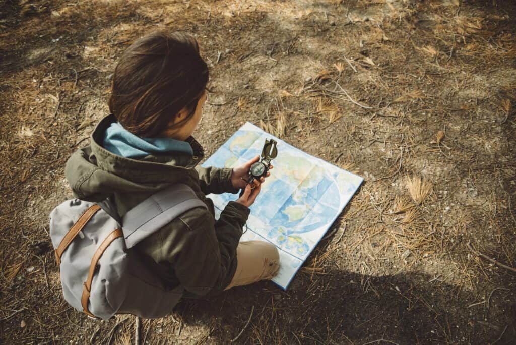 A hiker crouches on dry forest ground, looking at a compass in her hands while a world map lies open on the soil beside her — a metaphor for the DIKW model: the map defines the levels, but orientation requires judgement the map cannot provide.